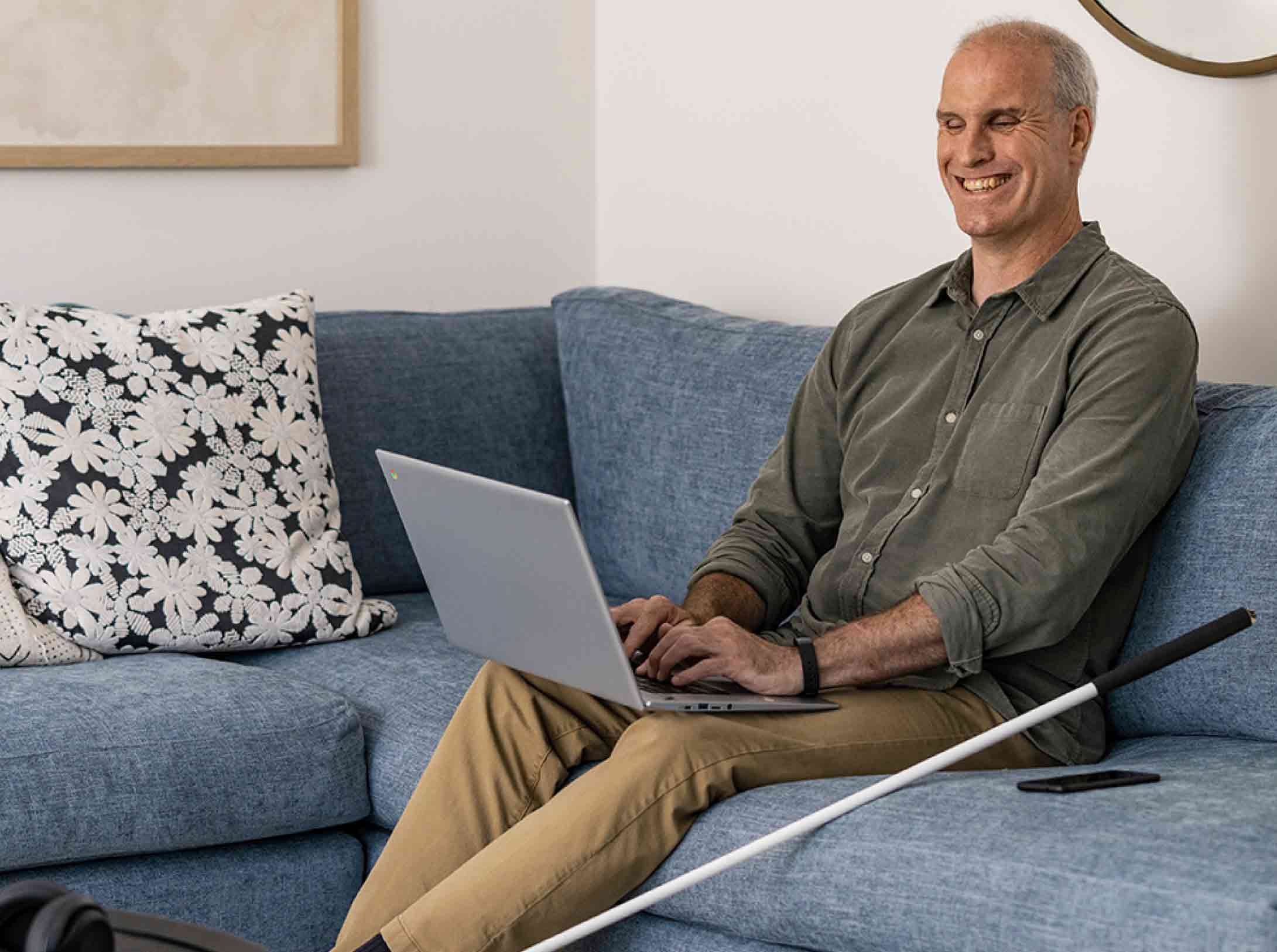 Een ouder persoon zit op een blauwe bank te typen op een geopende Chromebook op schoot terwijl aan diens linkerkant een blindenstok tegen de bank leunt.