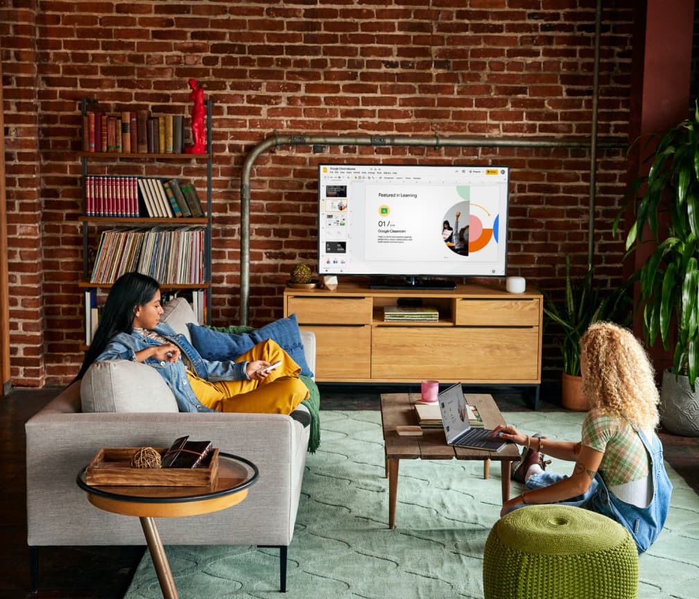 Iemand zit op een bank naar een tv-presentatie te kijken. Een tweede persoon zit op de grond aan een koffietafel met een Chromebook.